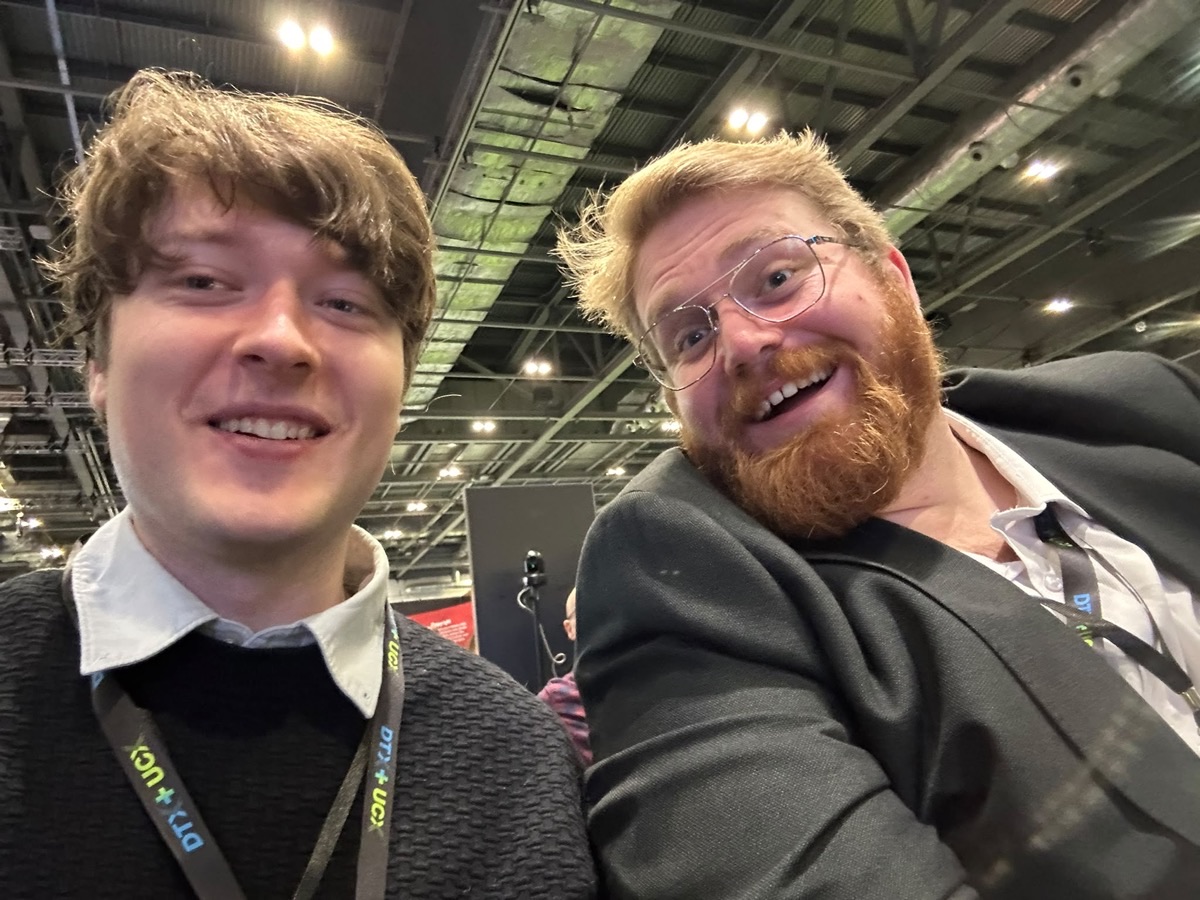 Dan and Jonathan smiling for a selfie in the exhibition hall, both wearing DTX + UCX lanyards